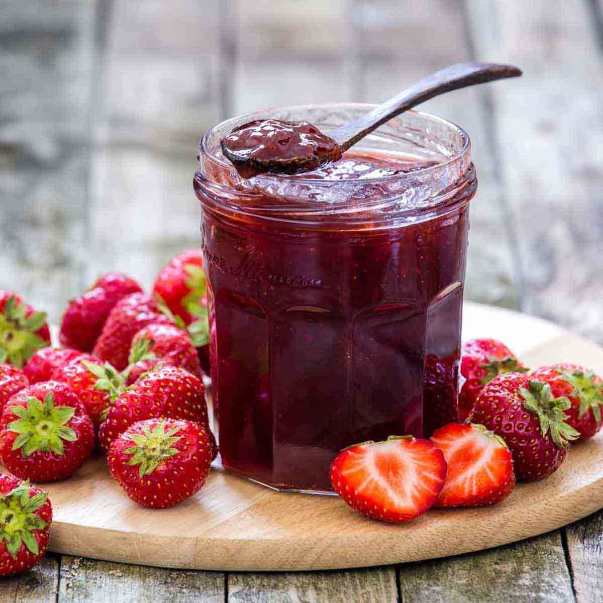 A jar of strawberry jam with a spoon rests on a wooden board, surrounded by fresh strawberries, showcasing vibrant red colors and a glossy texture.