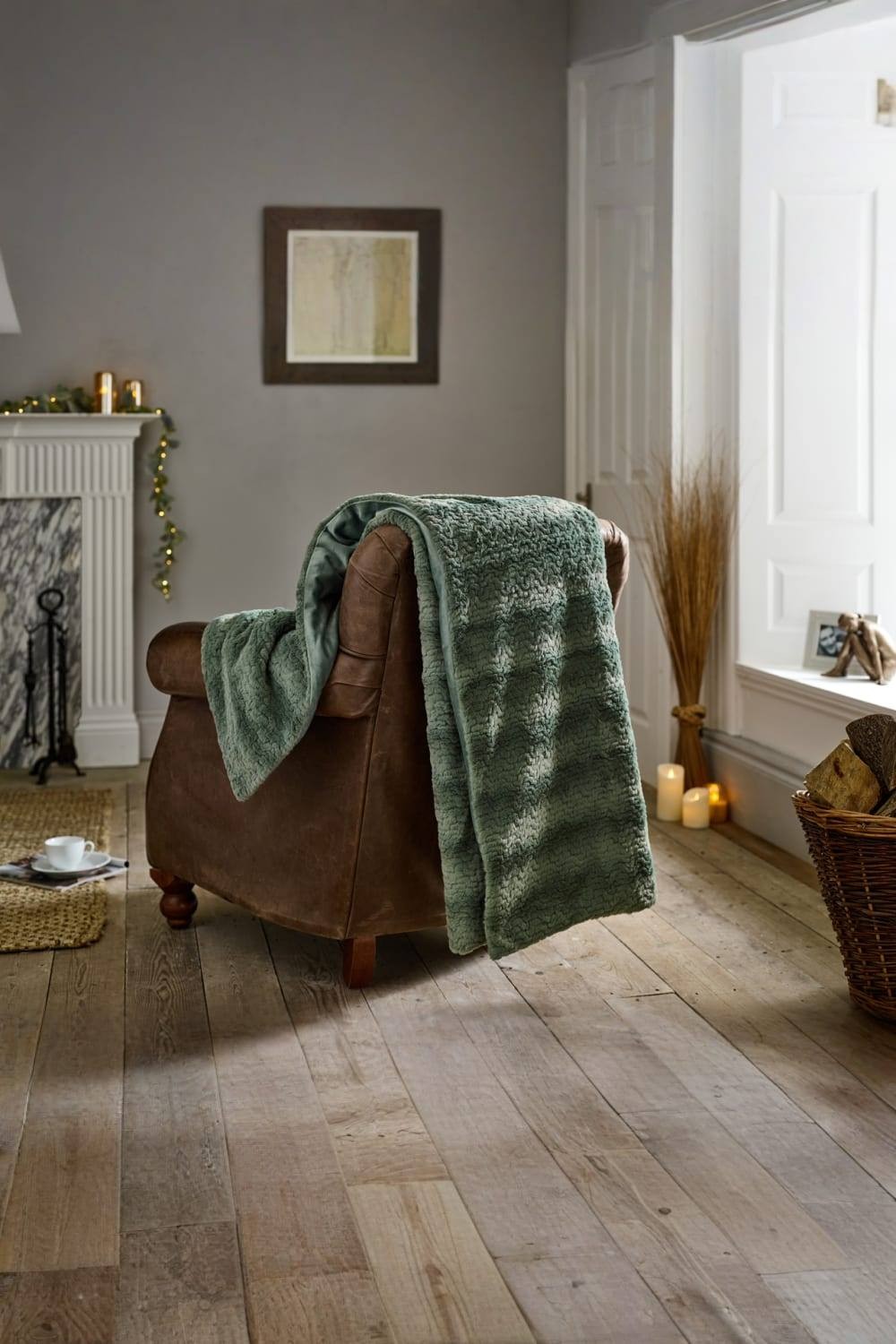 Soft green faux fur throw measuring 130x170cm draped over a brown leather armchair, set in a cozy room with wooden flooring and warm lighting.