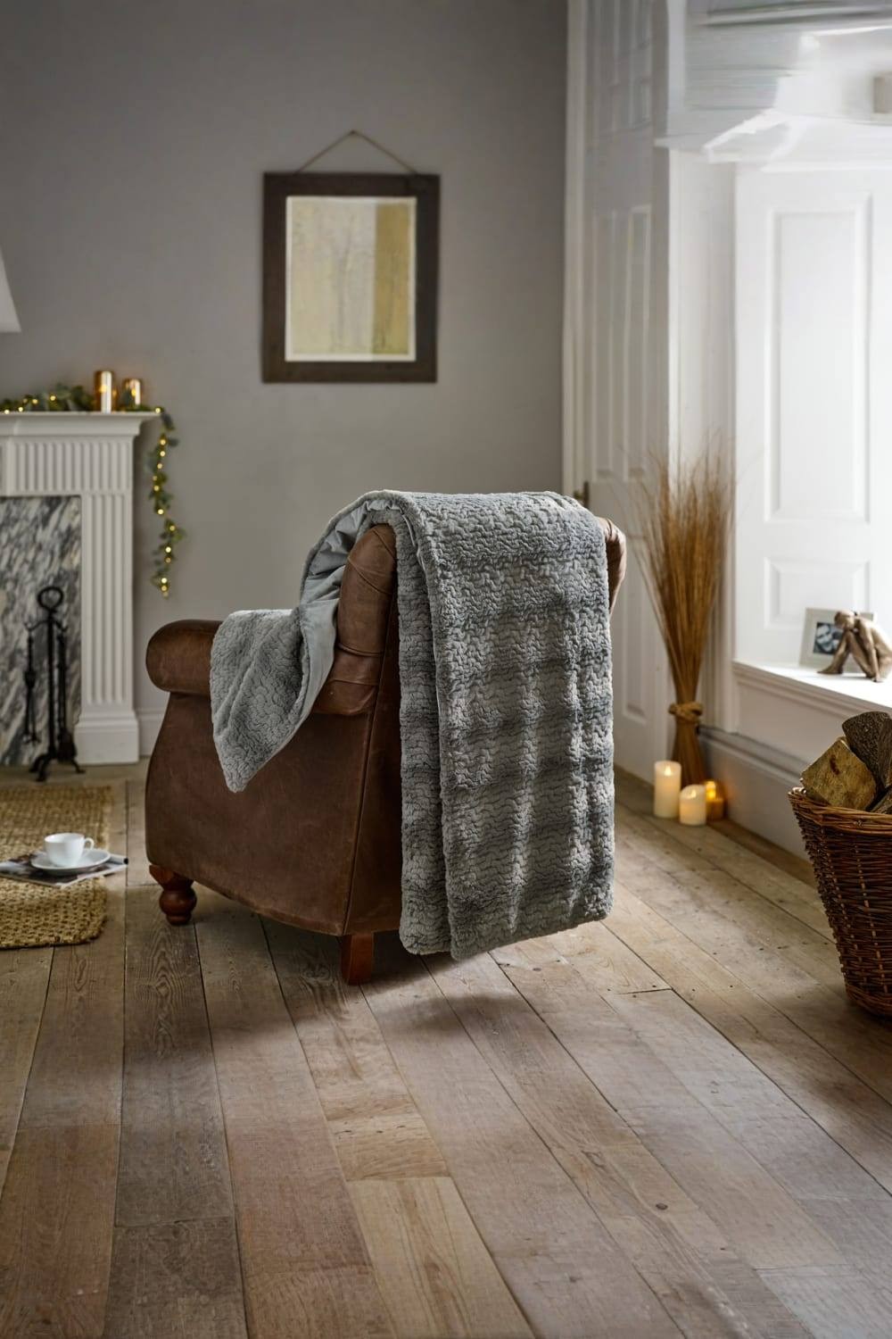 Soft grey faux fur throw measuring 130x170cm draped over a brown leather armchair, with a cozy living room setting featuring a fireplace and decorative elements.