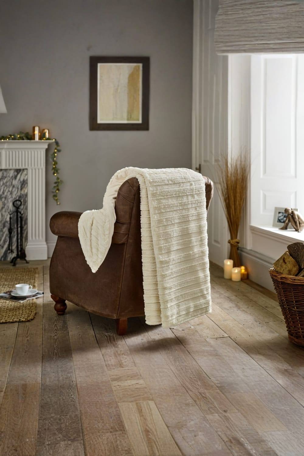 Cream-colored soft faux fur throw draped over a brown leather armchair, with a cozy interior setting featuring candles and decorative elements in a warm, inviting room.