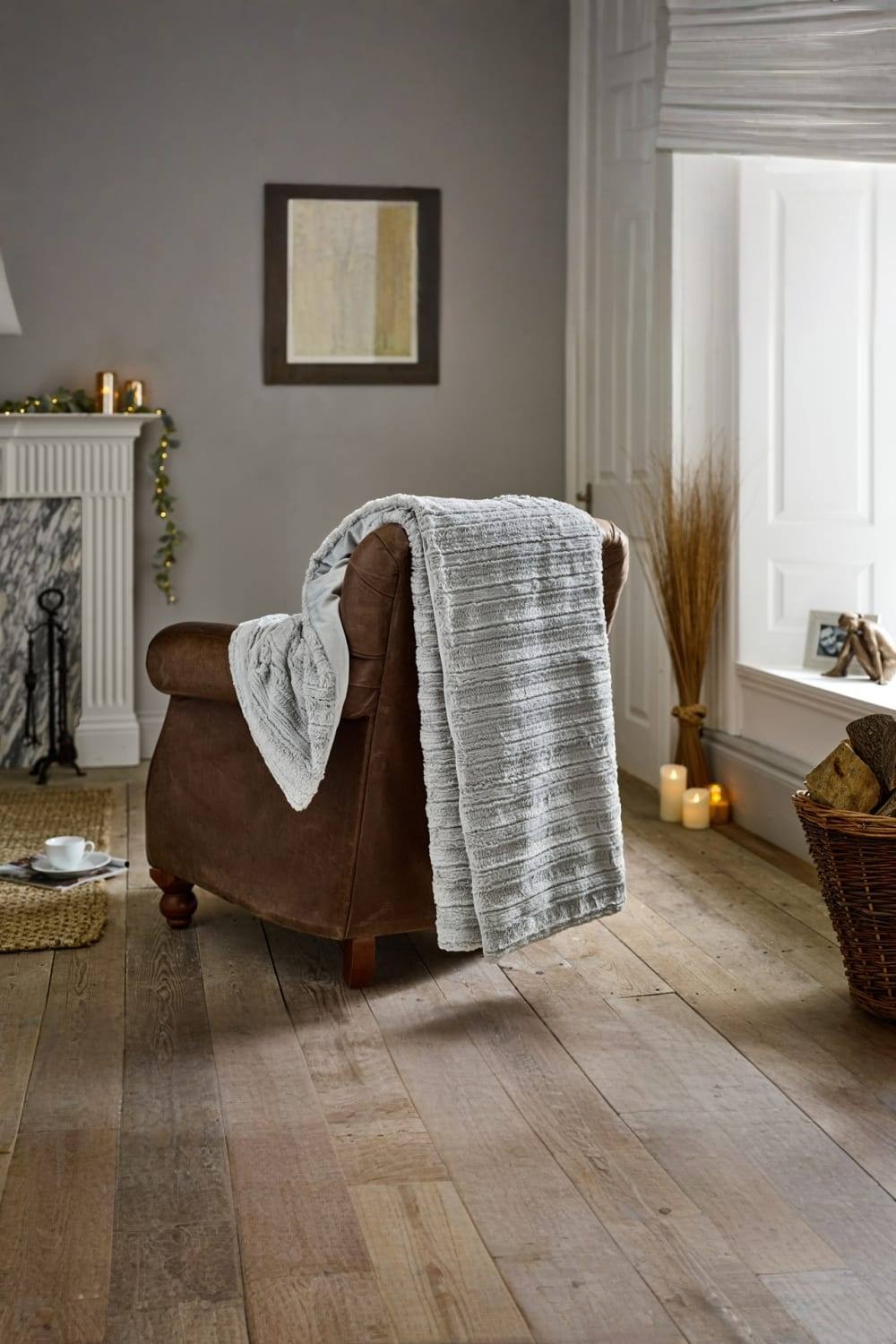 Soft grey faux fur throw draped over a brown leather armchair, featuring a textured design, measuring 130x170cm, set in a cozy, warmly lit room.
