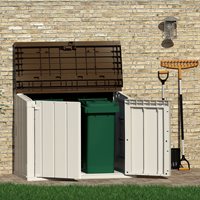 Extra large grey plastic outdoor garden storage unit with double doors, housing a green wheelie bin, set against a stone wall with a rake and shovel nearby.