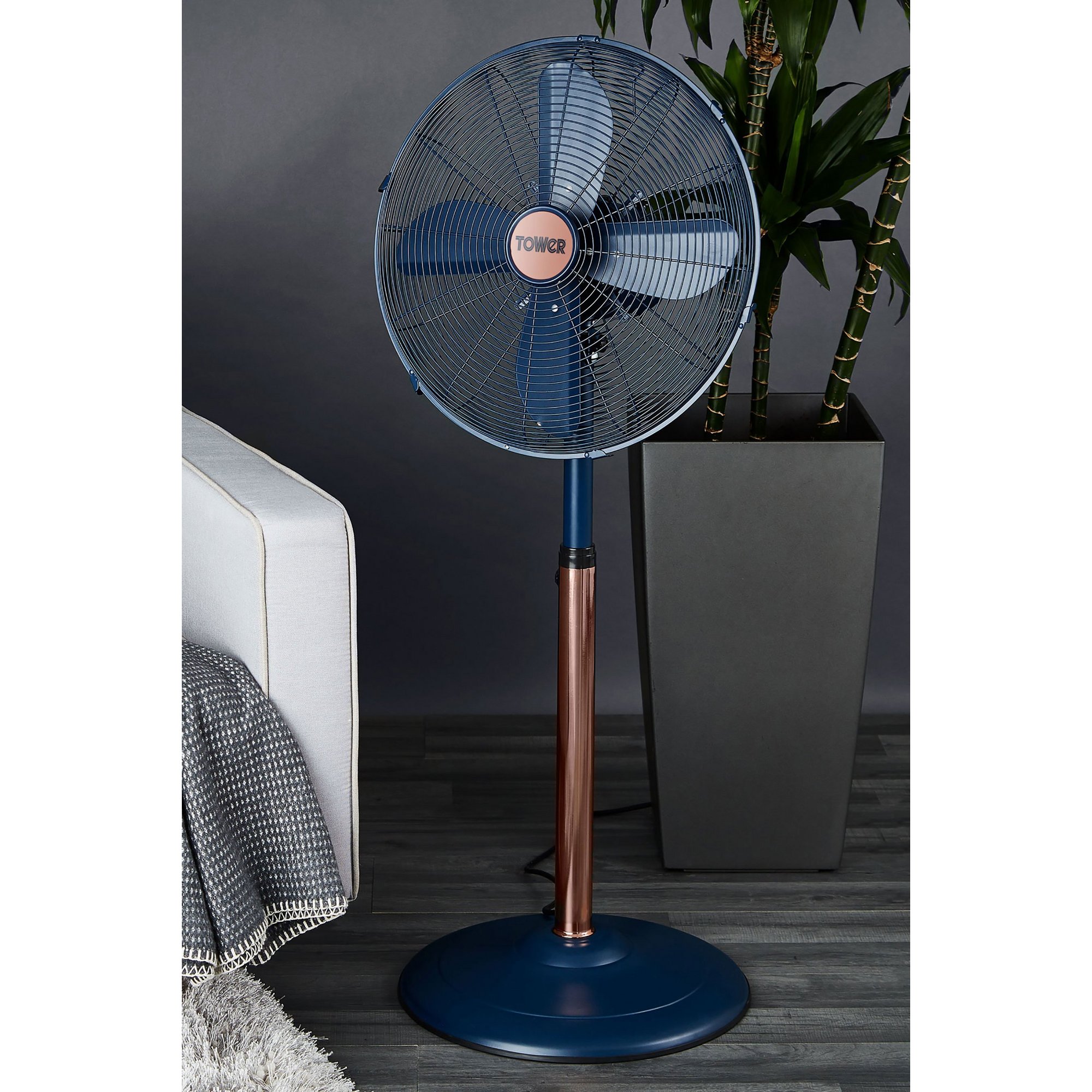 Midnight blue 16-inch pedestal fan with a copper-toned pole, featuring a protective metal grille and five blades, positioned beside a potted plant and sofa.