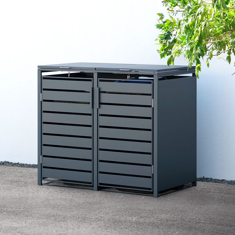 Anthracite metal wheelie bin store featuring double compartments, slatted design, and a flat top, measuring 1.32m x 0.80m, set against a light wall and greenery.