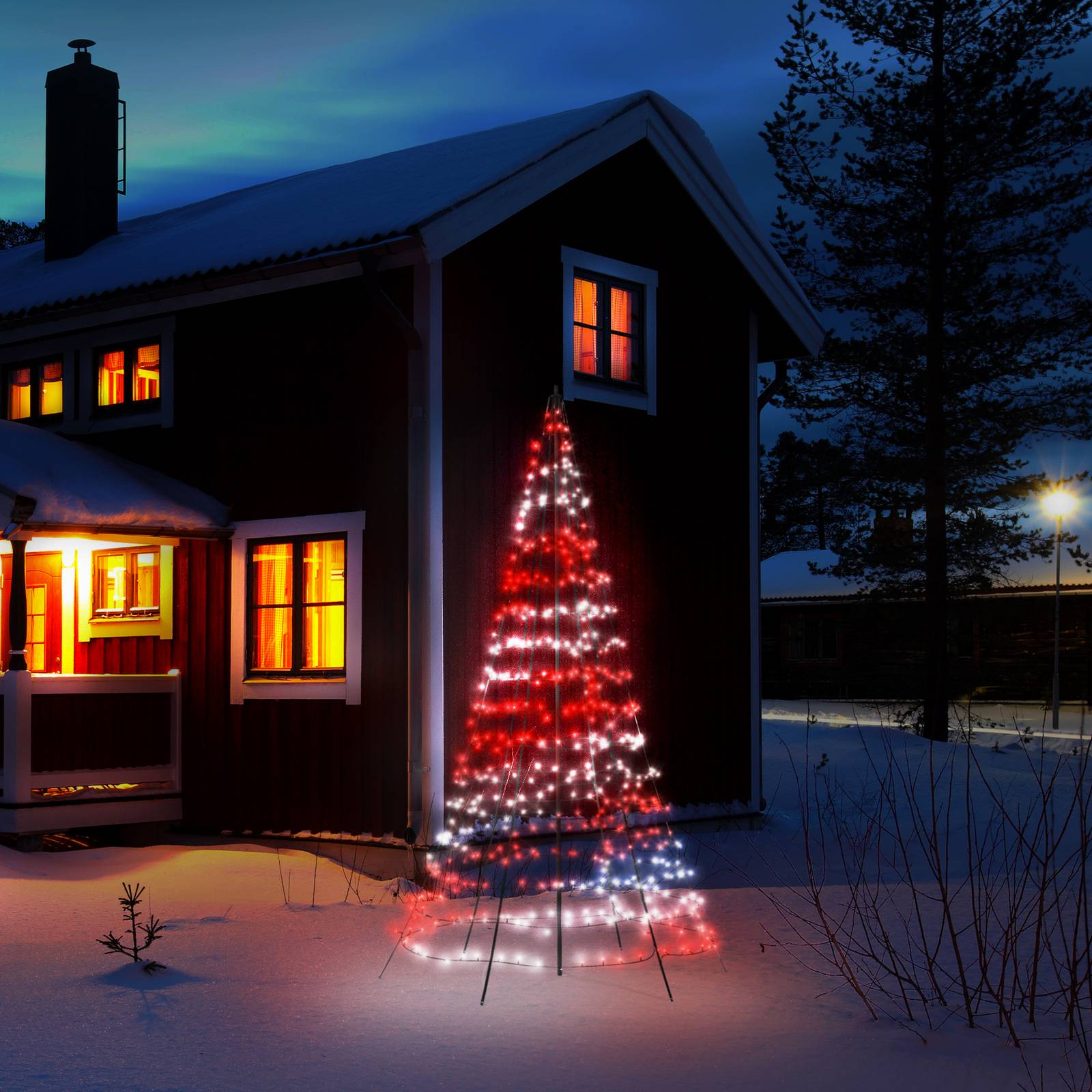 A tall, 300cm outdoor light tree in red and white RGBW colors, set against a snowy backdrop near a cozy cabin with warm glowing windows.