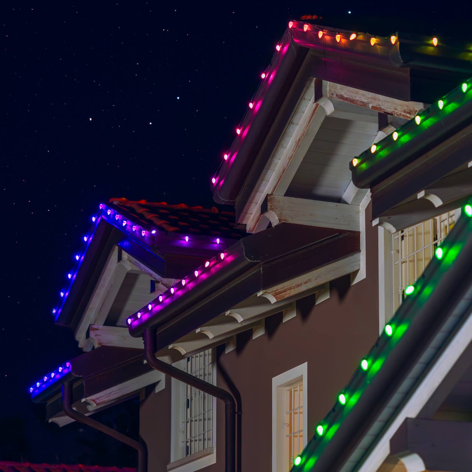 Colorful TWINKLY C9 Pine Cone Generation II Smart RGB LED string lights in purple, blue, and green adorn the edges of a house roof against a starry night sky.
