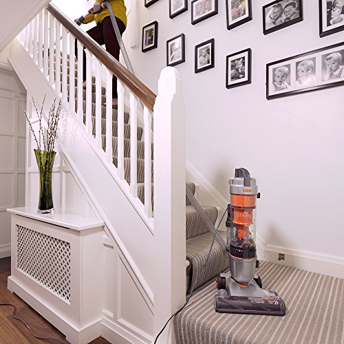 Vax U85-AS-BE upright corded bagless vacuum in orange and grey, featuring a transparent dust container, a sleek design, and a long power cord, positioned on a carpeted staircase.