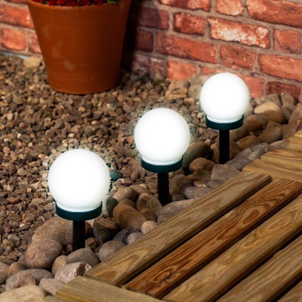 Three white frosted globe solar-powered outdoor spike lights with black bases, arranged along a stone pathway beside a wooden deck and a potted plant in the background.