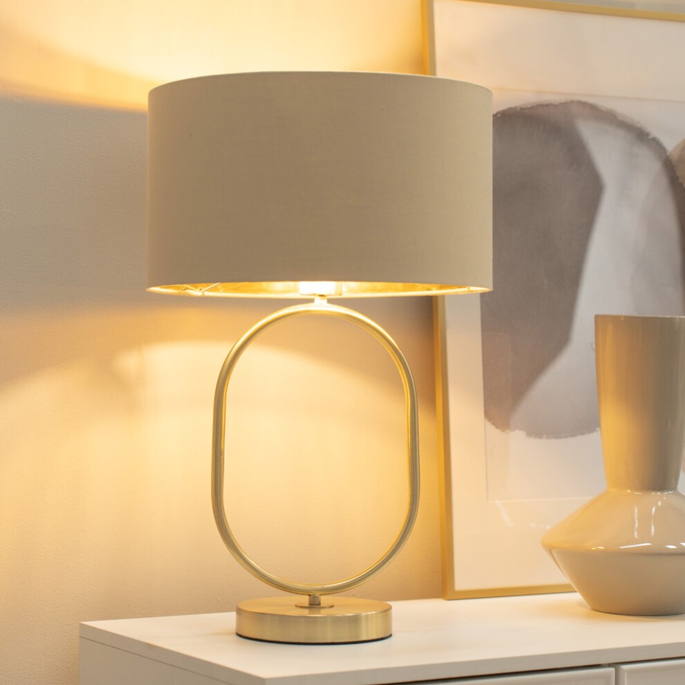 Brushed gold oval table lamp with a beige fabric drum shade, featuring a sleek design and an LED bulb, set against a soft, warm background.