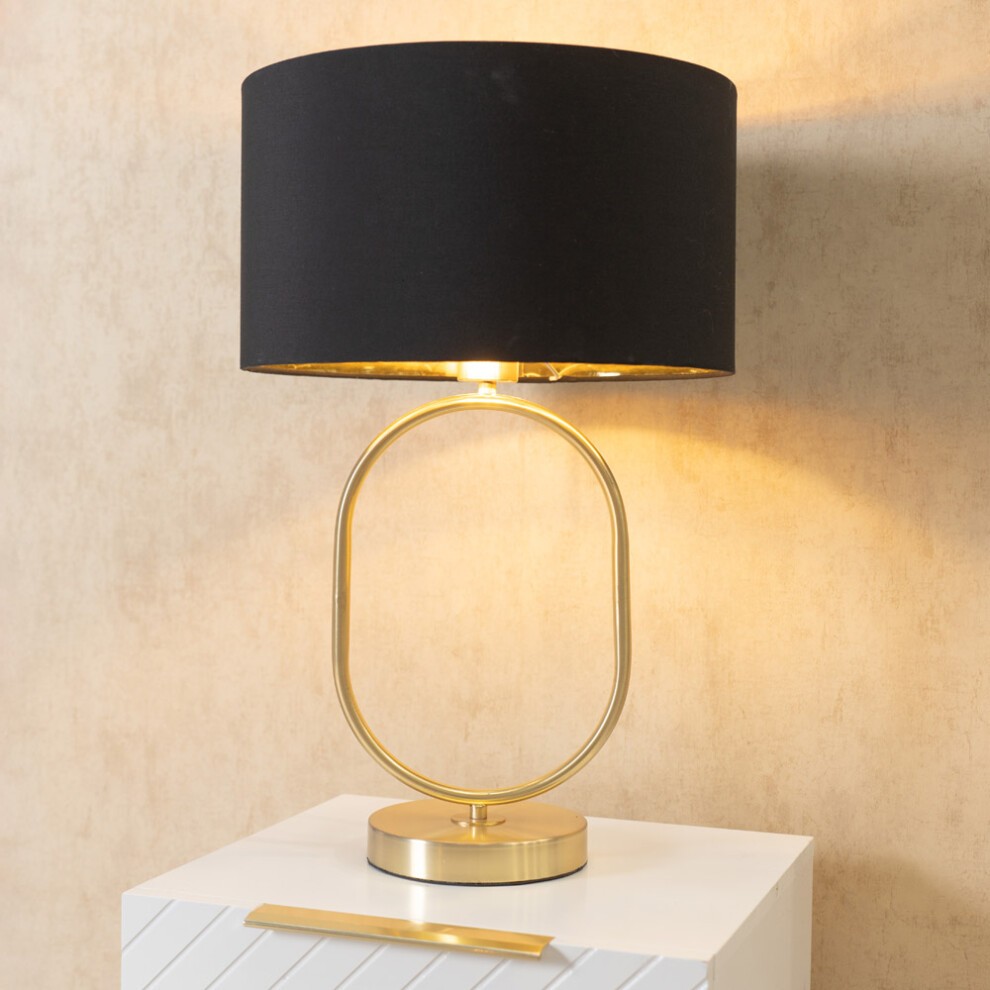 Brushed gold oval table lamp featuring a black fabric drum shade, illuminated by an LED bulb, placed on a white textured surface.