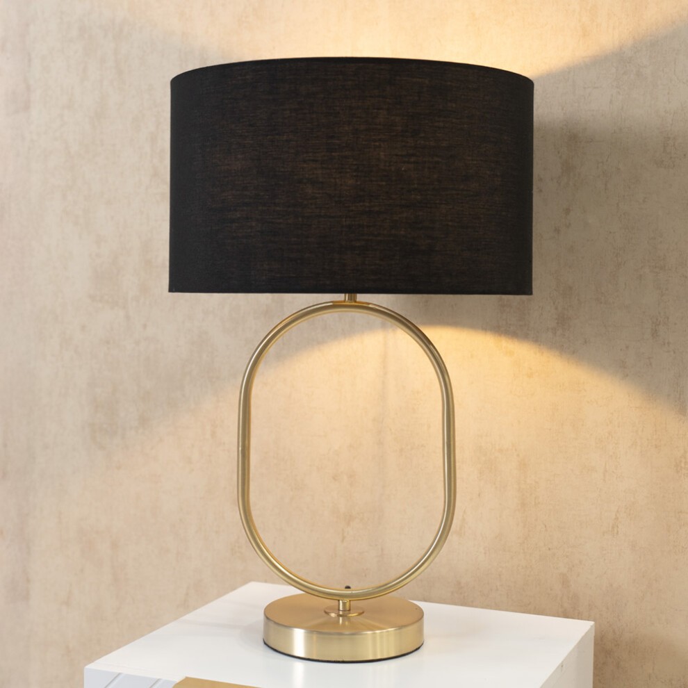 Brushed gold oval table lamp with a black fabric drum shade, featuring a sleek design and an LED bulb, set on a white side table against a textured beige wall.