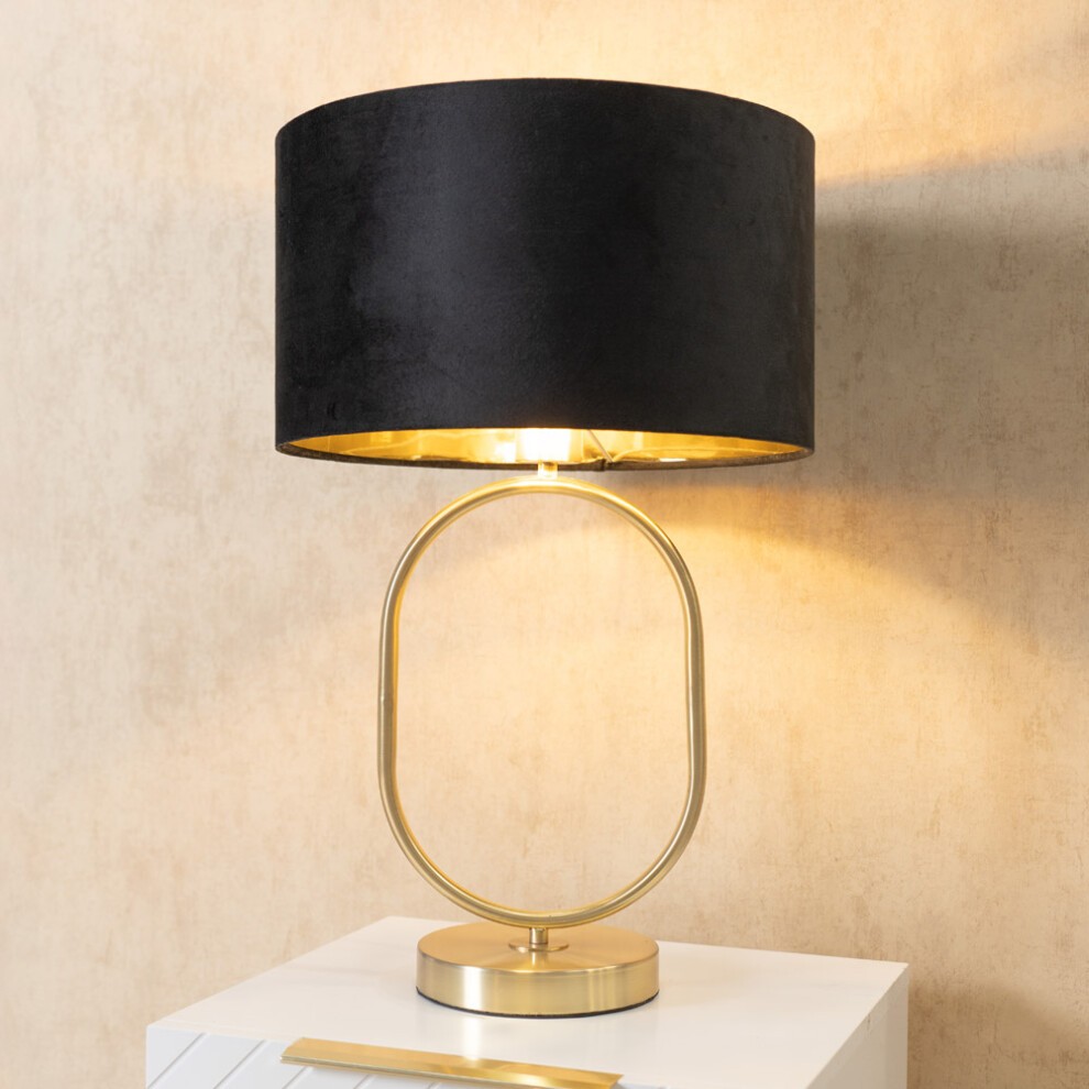 Black velvet drum shade with a brushed gold oval base and stand, featuring a warm LED bulb, elegantly illuminating a textured wall.