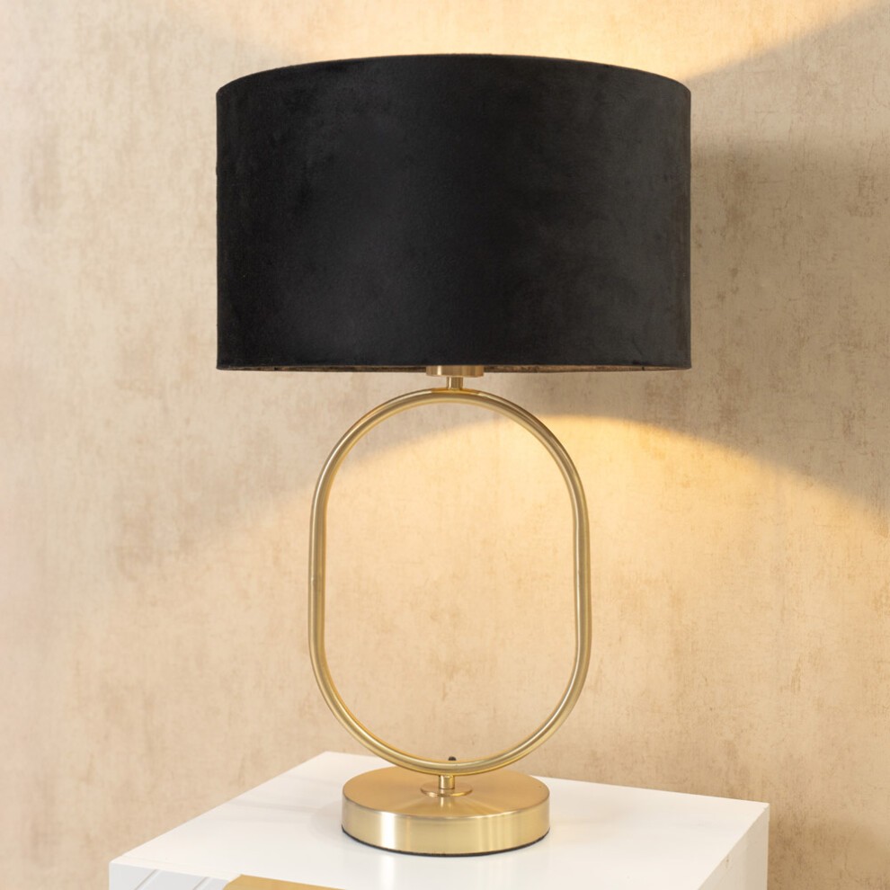 Black velvet drum lamp shade atop a brushed gold oval base, featuring a modern design and includes an LED bulb, casting a warm glow against a textured background.