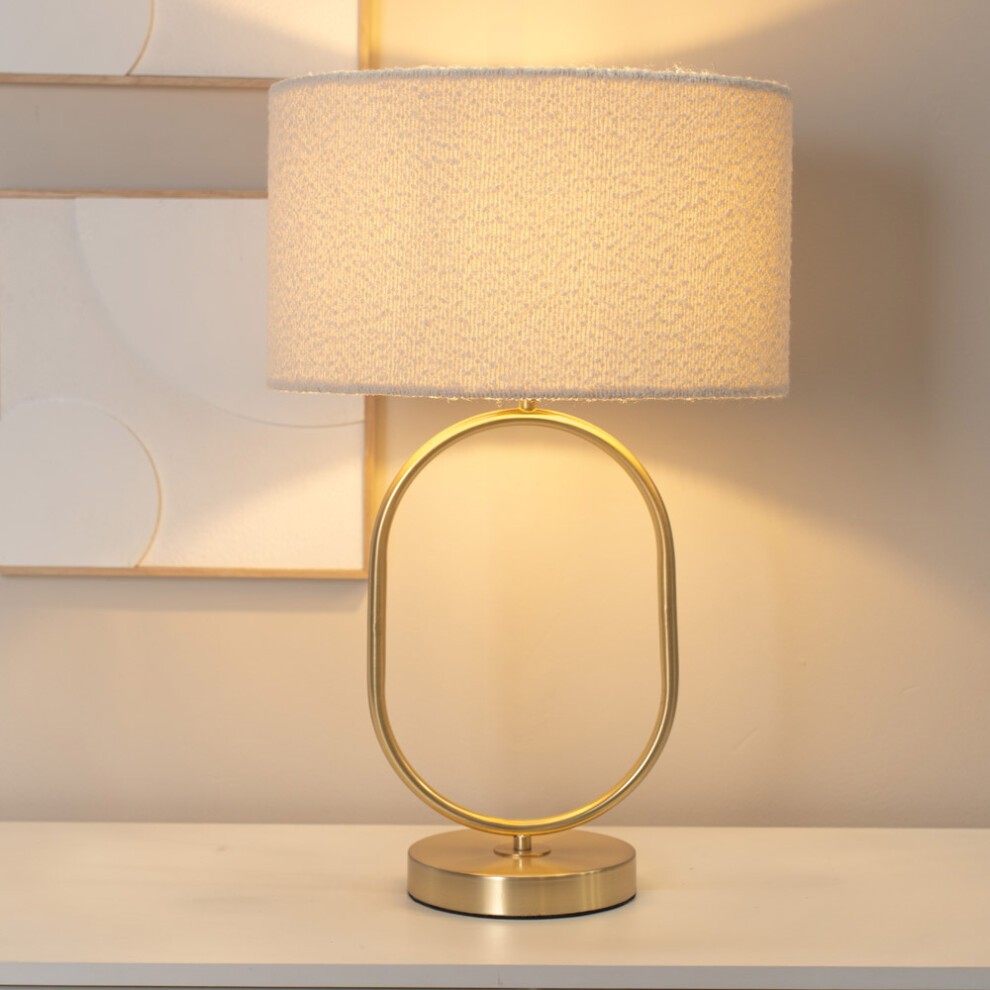 Antonia cream boucle drum shade table lamp with a brushed gold oval base, featuring a modern design and an included LED bulb, casting a warm glow.