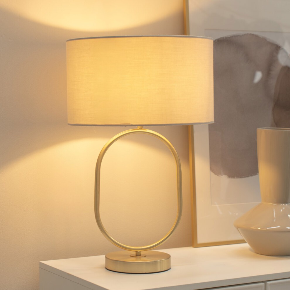 Brushed gold oval table lamp with a natural fabric drum shade, featuring a sleek circular base and warm LED light, set against a neutral background.