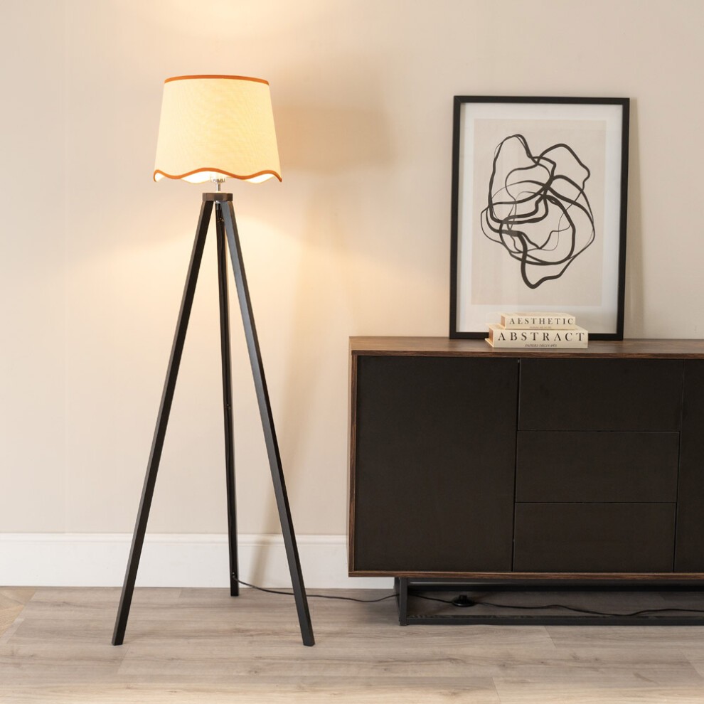 Black wood tripod floor lamp featuring a scallop edge linen shade and an LED bulb, positioned beside a dark cabinet and an abstract framed artwork.