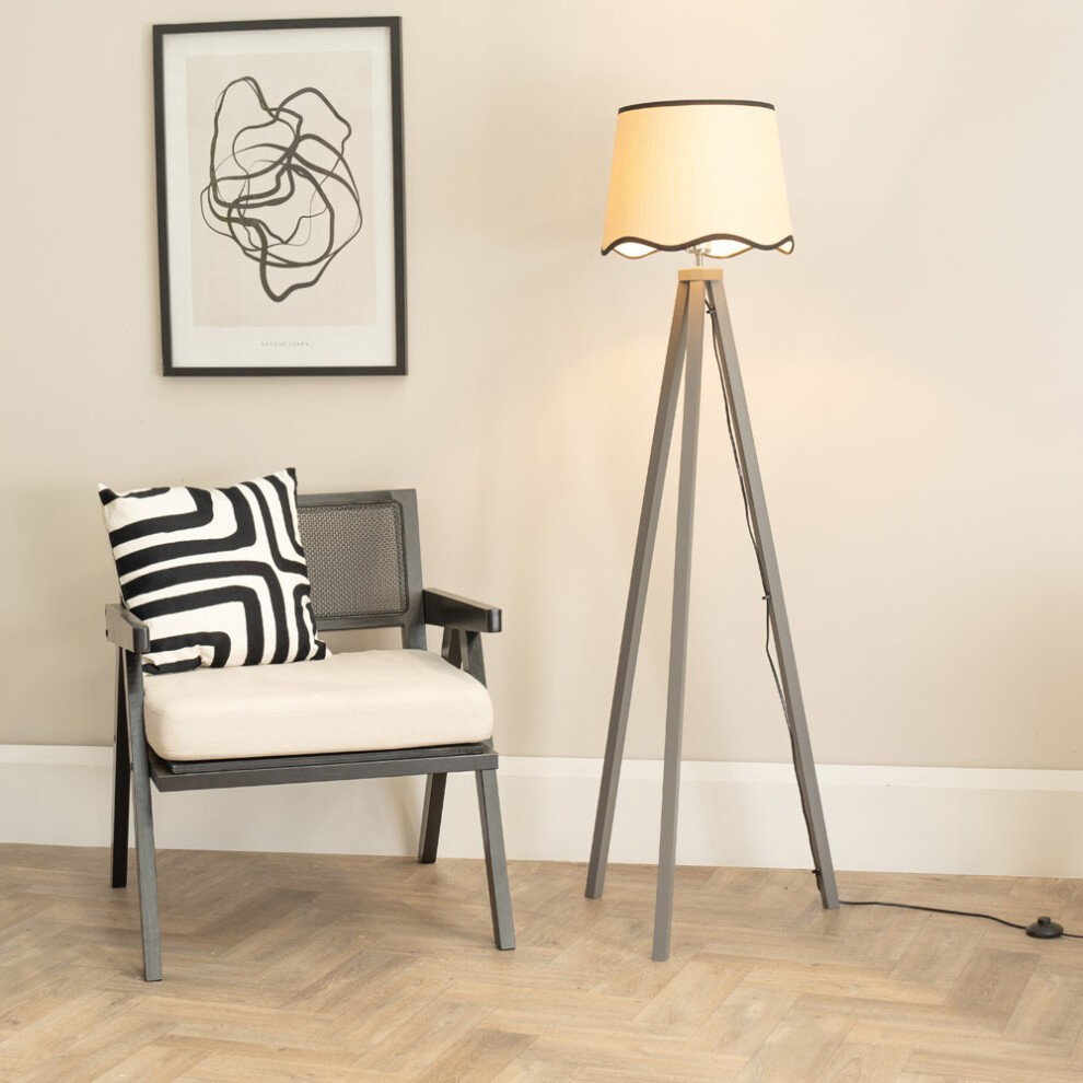 Grey wood tripod floor lamp with a scallop-edged linen shade, accompanied by a black and white striped cushion on a modern chair against a neutral wall.