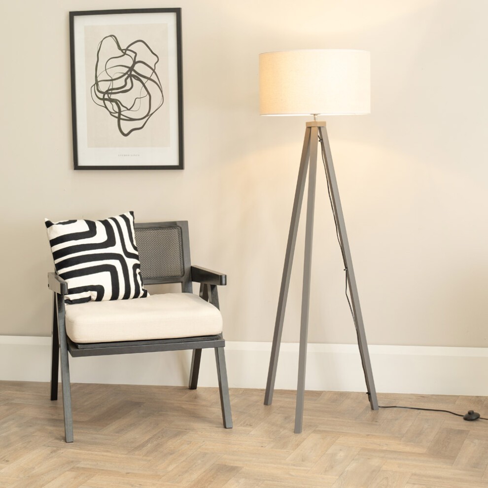 Grey wood tripod floor lamp with a beige linen drum shade, accompanied by a black and white striped cushion on a modern chair, set against a neutral wall.