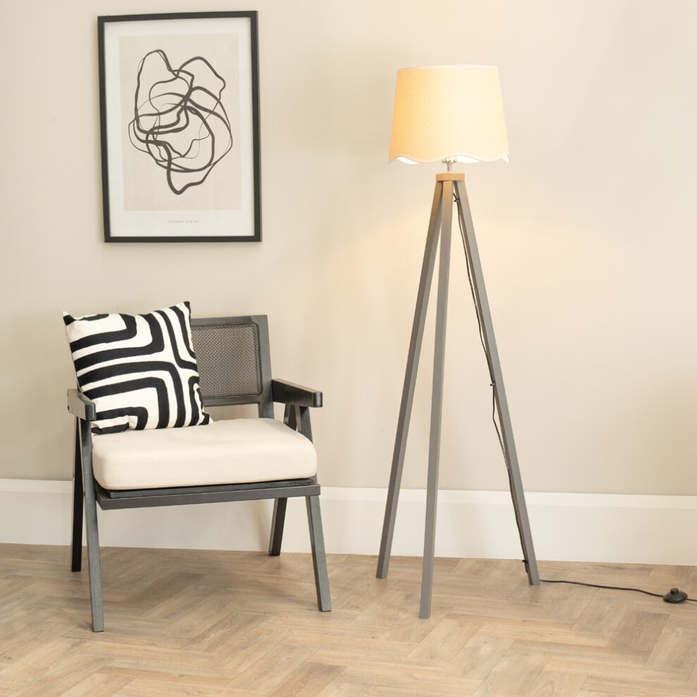 Grey wood tripod floor lamp with a scalloped linen shade, accompanied by a black and white striped cushion on a modern chair, set against a neutral wall with abstract art.
