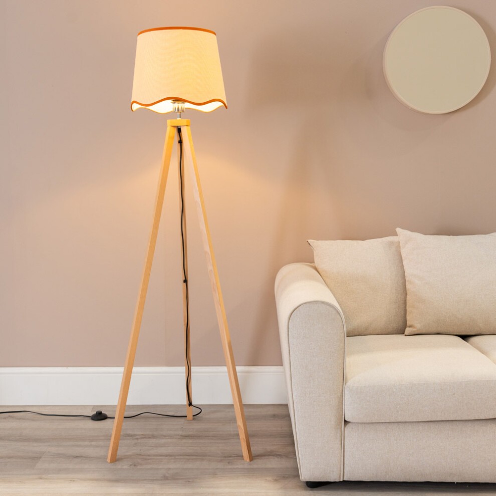 Light wood tripod floor lamp with a scallop edge linen shade, featuring an LED bulb, positioned beside a beige sofa against a soft pink wall.