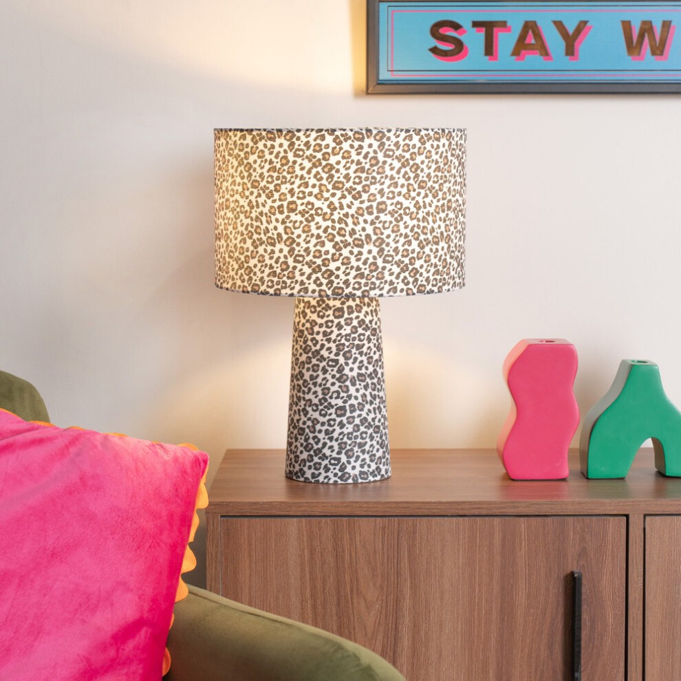 Tan leopard print fabric table lamp with a cylindrical base and a wide shade, illuminated, placed on a wooden table beside colorful decor items.