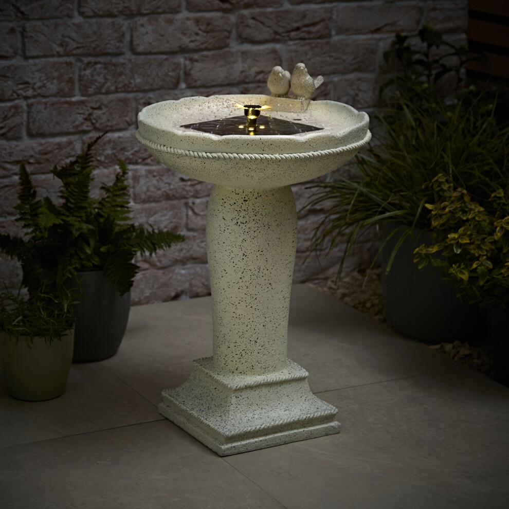 Solar-powered bird bath fountain in white, featuring a textured bowl and a pedestal base, with two decorative birds perched on the rim, surrounded by greenery.
