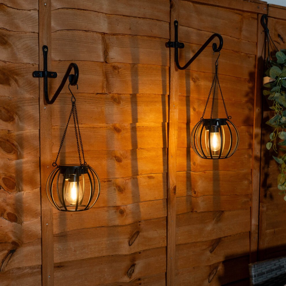 Two black metal ball lanterns hang from hooks on a wooden wall, featuring a warm glow from the solar-powered bulbs inside, perfect for outdoor garden decor.