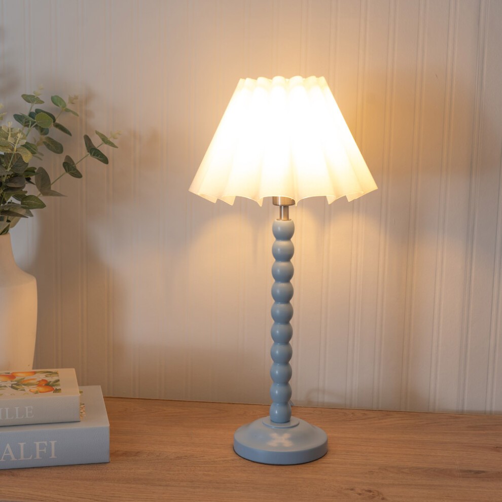 Powder blue table lamp with a scallop tapered white shade and LED bulb, featuring a beaded design on the stem, set against a wooden surface with greenery and books.