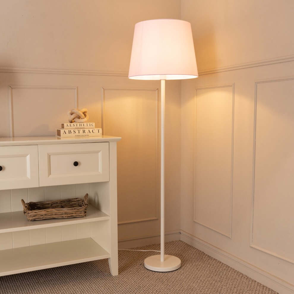White single stem floor lamp with a pink fabric tapered shade, standing beside a white cabinet with a decorative sign and a woven basket on a light-colored carpet.