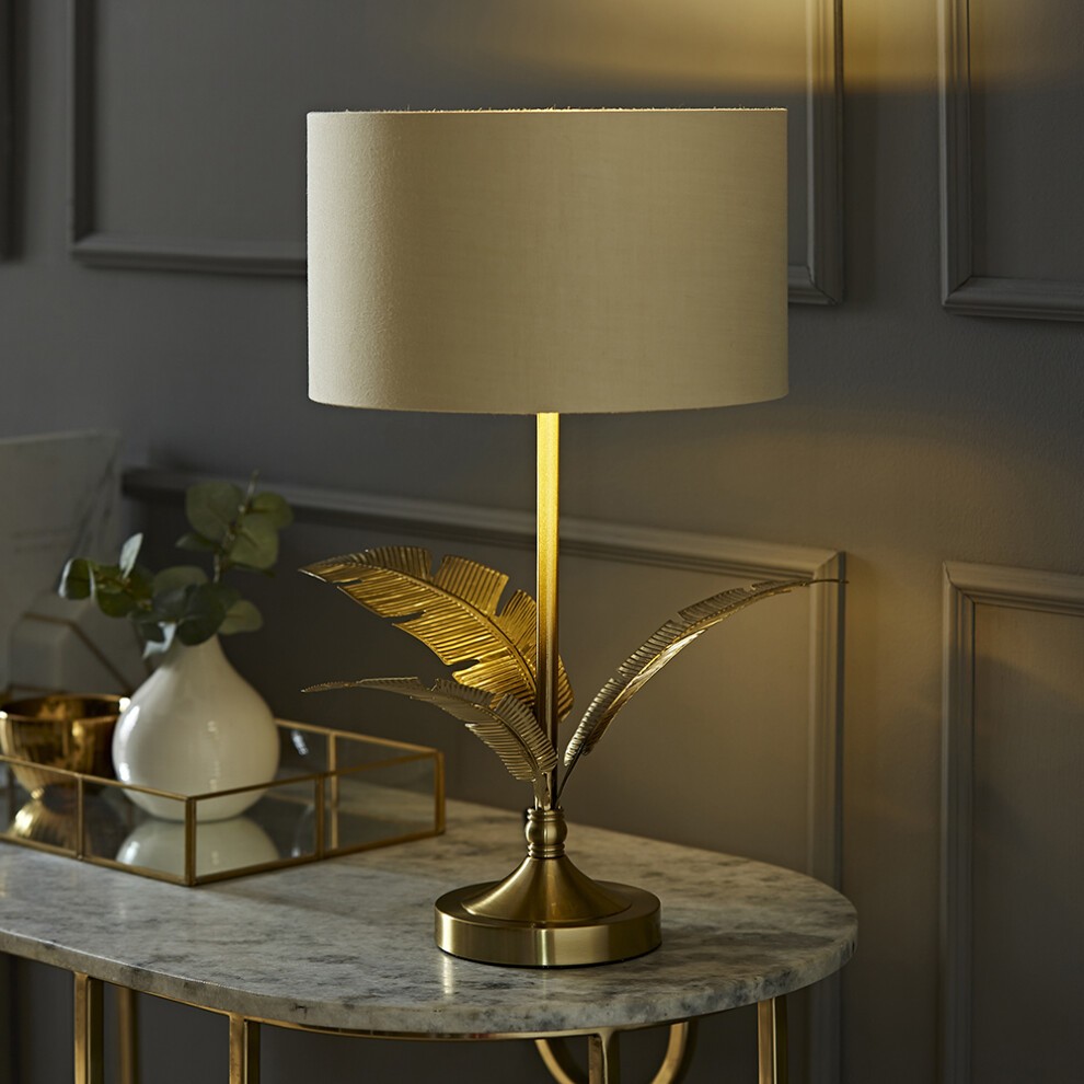 Gold palm leaf table lamp featuring a natural fabric drum shade, set on a sleek gold base, with a marble tabletop and decorative accessories in the background.