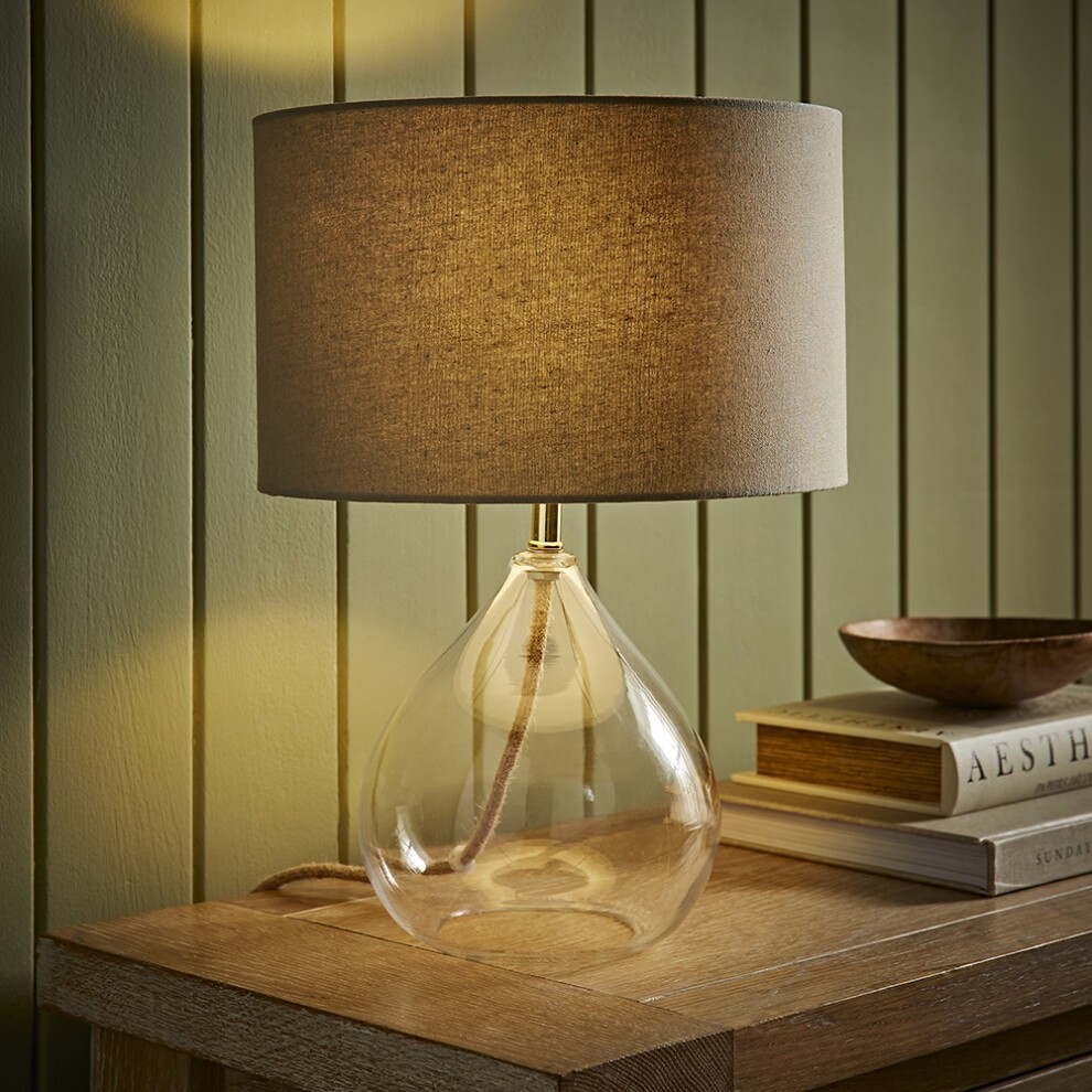 Natural linen drum shade table lamp with a clear glass teardrop base, featuring a gold accent and a warm, soft glow, placed on a wooden table next to stacked books.