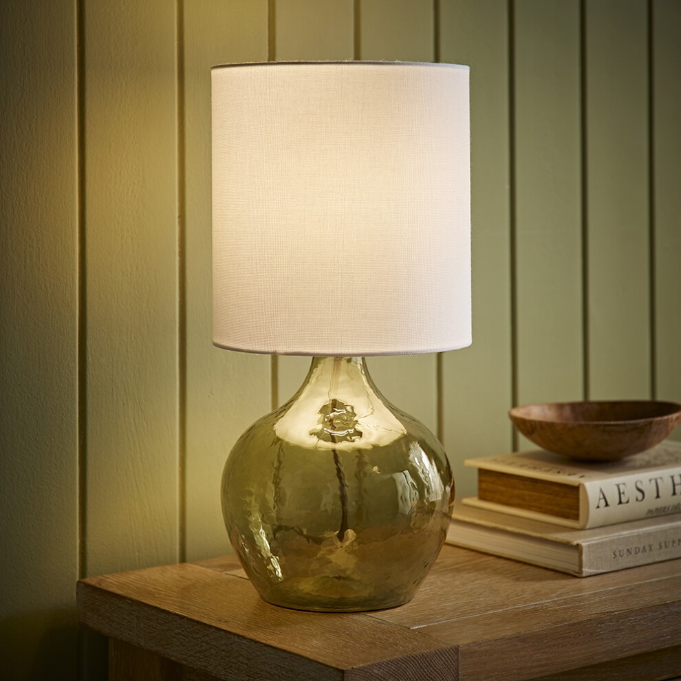 Natural linen drum shade atop a dark green glass teardrop base table lamp, featuring a warm glow, placed on a wooden surface with books and a bowl.