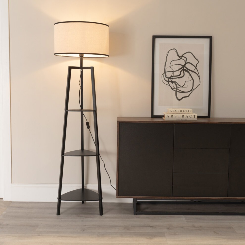 Black three-tier wood shelf floor lamp with a linen shade and LED bulb, standing next to a dark cabinet and framed abstract artwork on a light-colored wall.