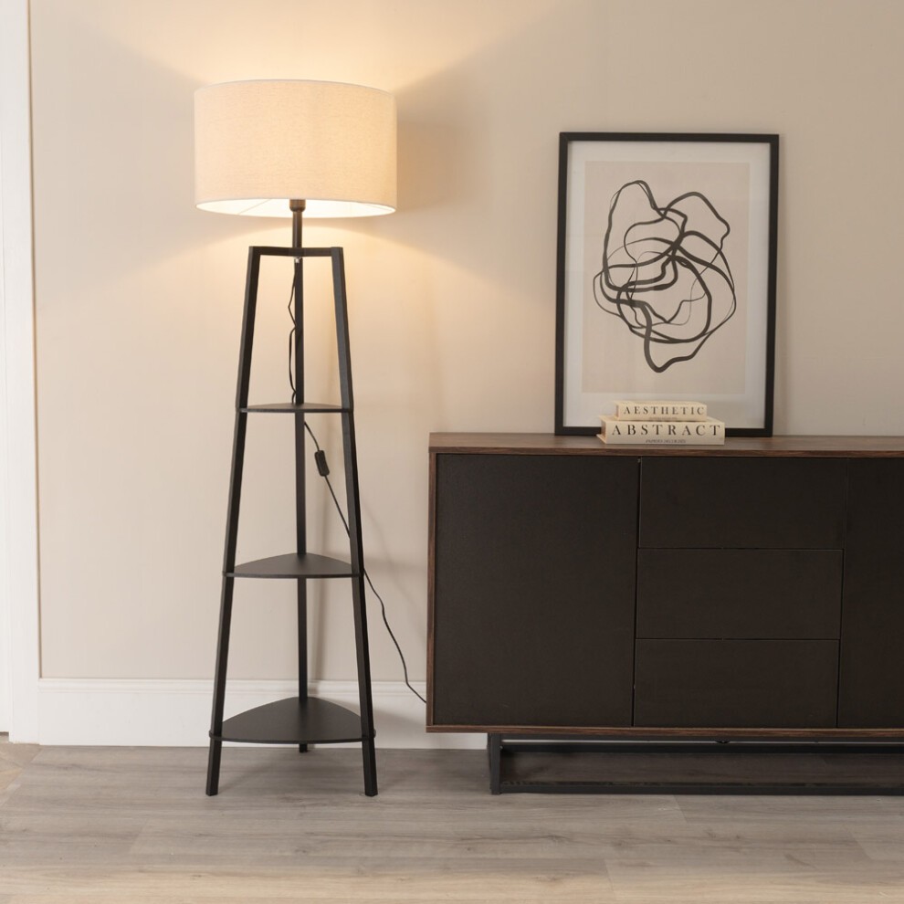 Black three-tier wood shelf floor lamp with a beige linen shade and included white LED bulb, positioned next to a dark cabinet and abstract art on the wall.