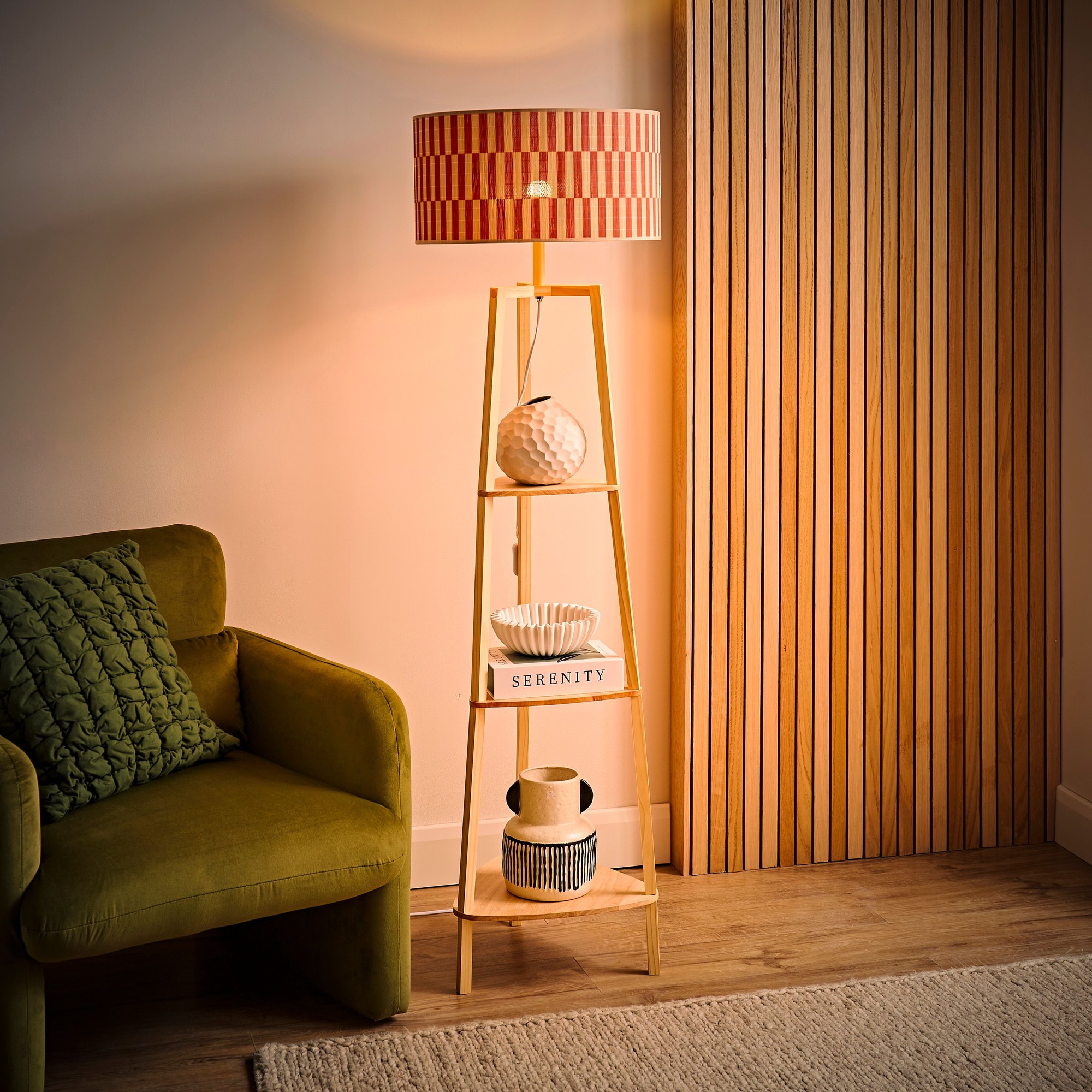 Hiru Shelf Tripod Floor Lamp features a bamboo frame with an orange checker patterned shade, illuminating a cozy space alongside decorative items on its shelves.