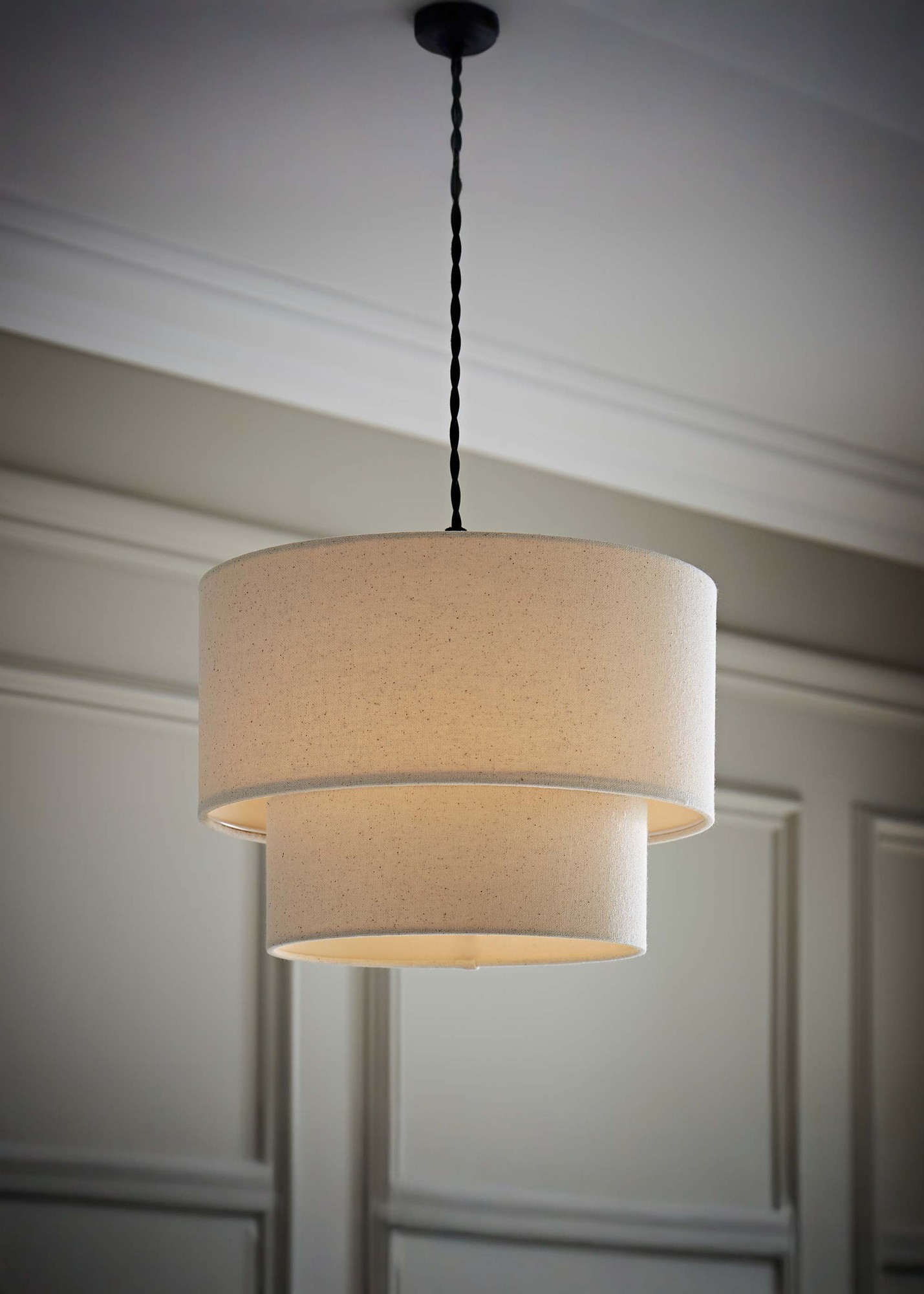 Natural linen two-tier drum pendant light with a soft beige shade, suspended by a black twisted cord, illuminating a stylish interior space.