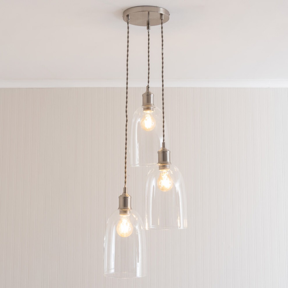Three glass cloche dome pendant lights hang from a silver ceiling mount, featuring LED bulbs and twisted fabric cords, set against a light-colored wall.