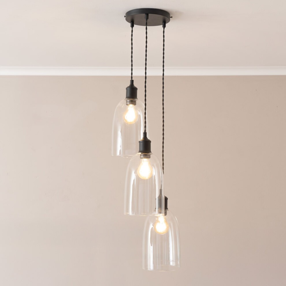 Three clear glass cloche pendant lights hang from a black ceiling mount, featuring LED bulbs and braided black cords, set against a neutral wall.