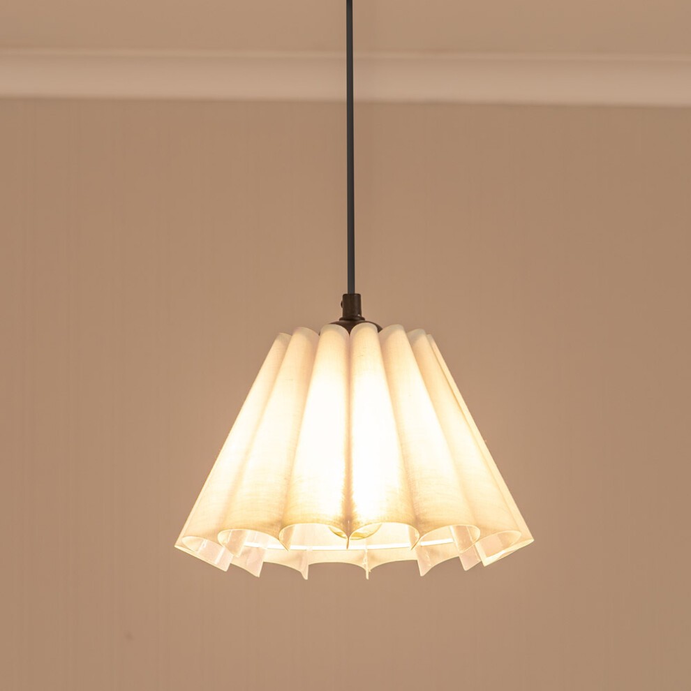 White fabric tapered lamp shade with pleated design and scalloped edge, suspended from a black cord, illuminating softly from within.