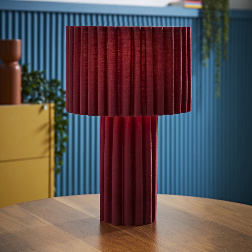 Burgundy ribbed fabric drum lamp shade atop a matching base, featuring a soft glow from the included bulb, set against a blue wall and wooden table.
