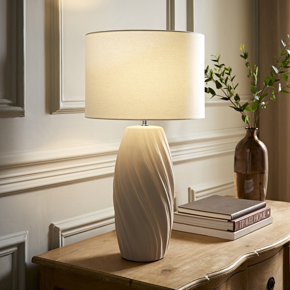Tall swirl-patterned table lamp with a natural linen drum shade, featuring a light beige ceramic base and an LED bulb, set on a wooden table beside stacked books and a vase.