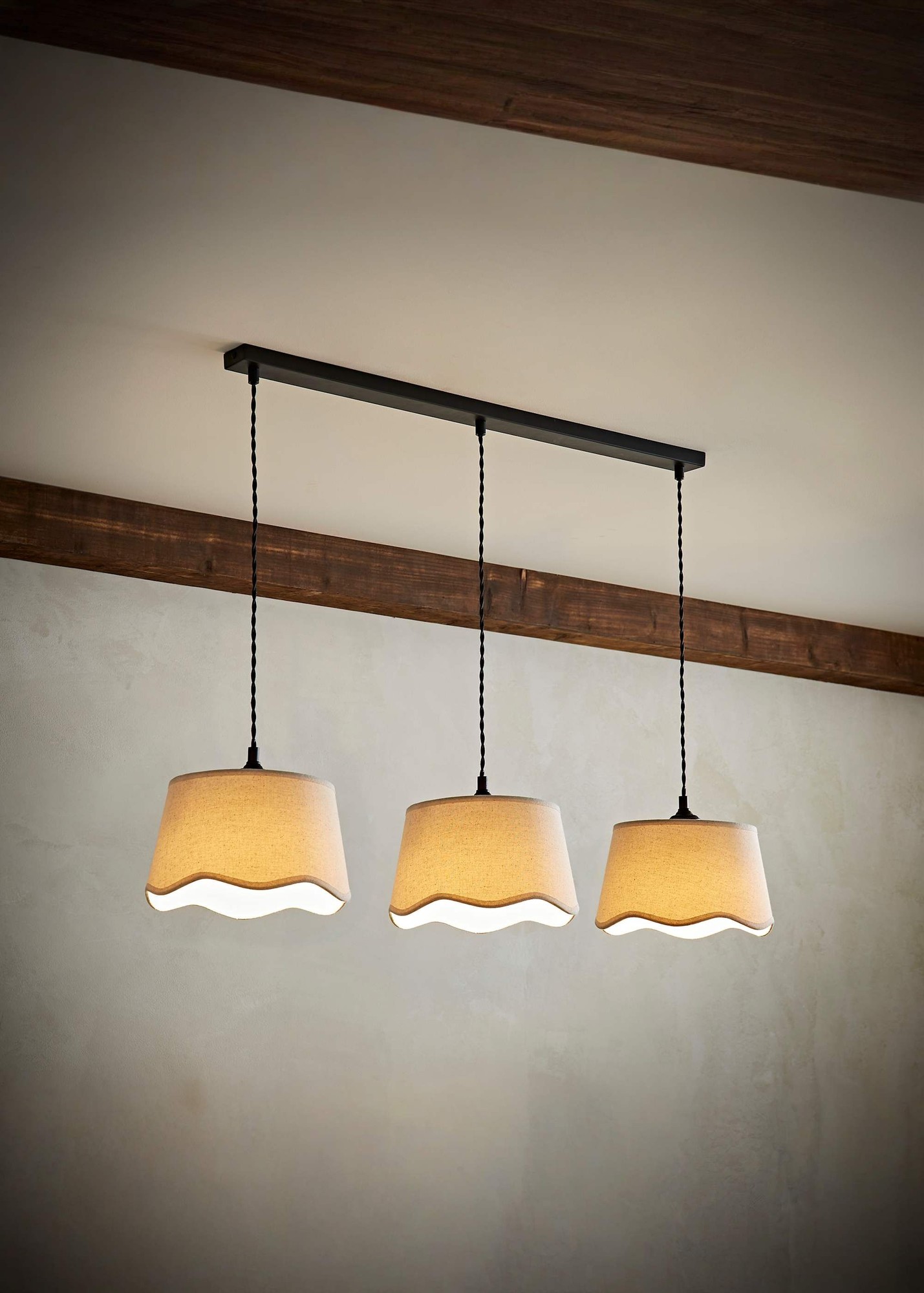Three scalloped natural linen lampshades in beige hang from a black metal bar, featuring a rustic wooden beam in the background, creating a warm, inviting ambiance.