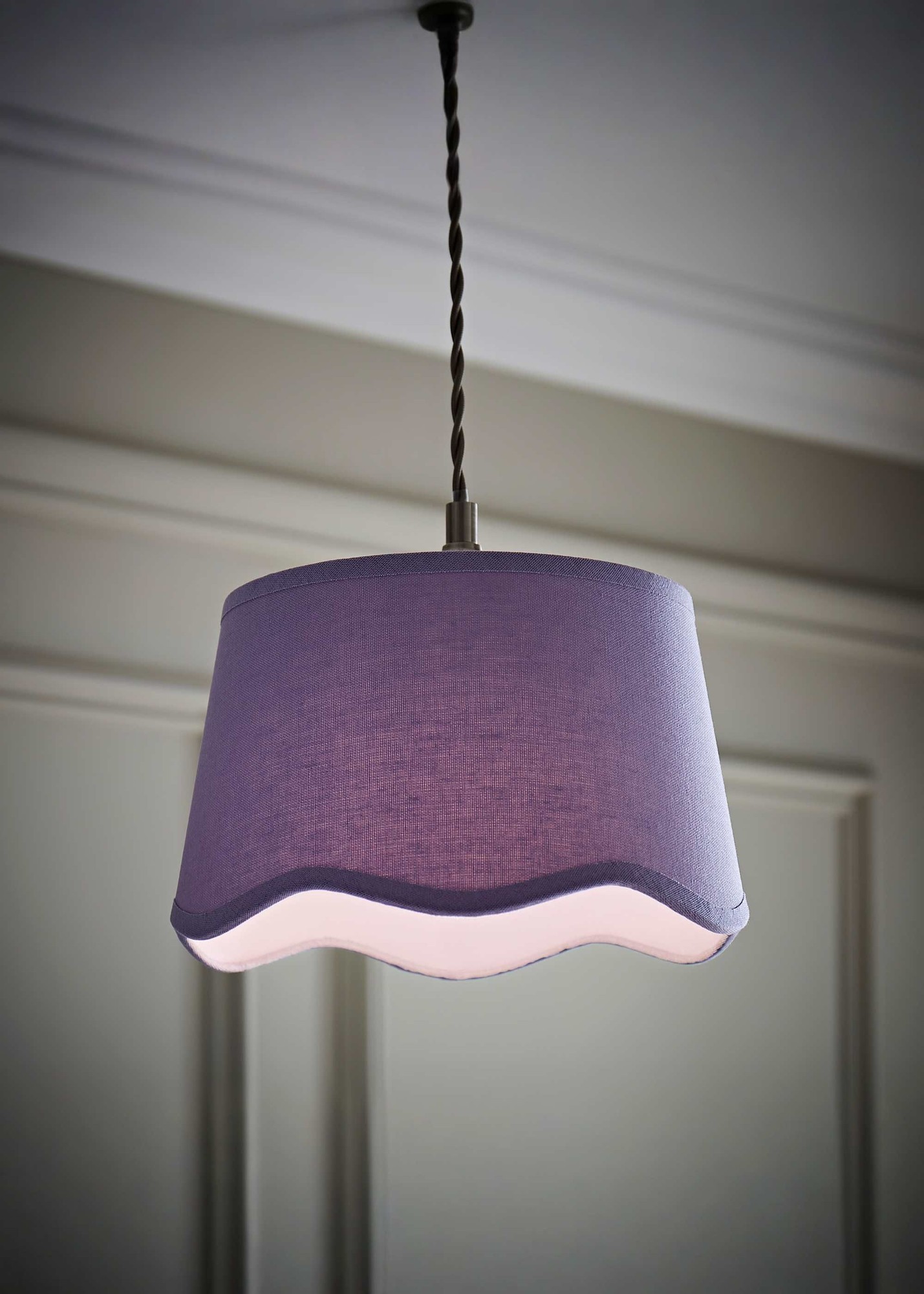 Small lilac ceiling lamp shade with a scalloped edge, made of linen, featuring a trim along the bottom, suspended from a twisted cord in a neutral setting.
