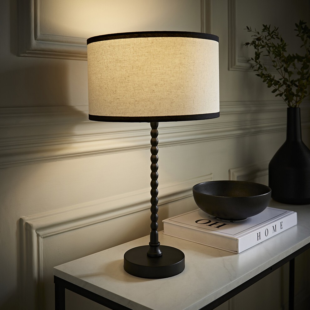 Natural linen drum shade with black trim atop a matte black bobbin table lamp, featuring a round base, set on a marble surface with a decorative bowl and books.
