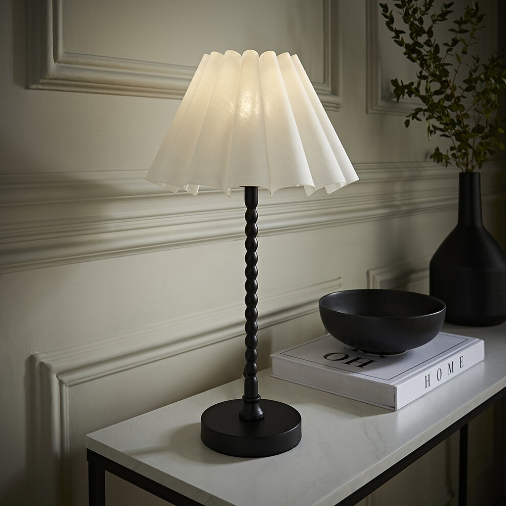 Matte black bobbin table lamp with a scallop pleated tapered shade in white, featuring a twisted base and positioned on a marble surface beside a decorative bowl and vase.