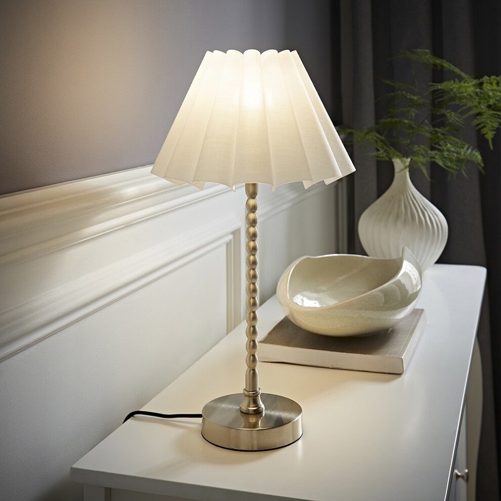 White scallop pleated taper shade atop a brushed chrome bobbin base table lamp, featuring a silver bulb, set on a light-colored surface with decorative items in the background.
