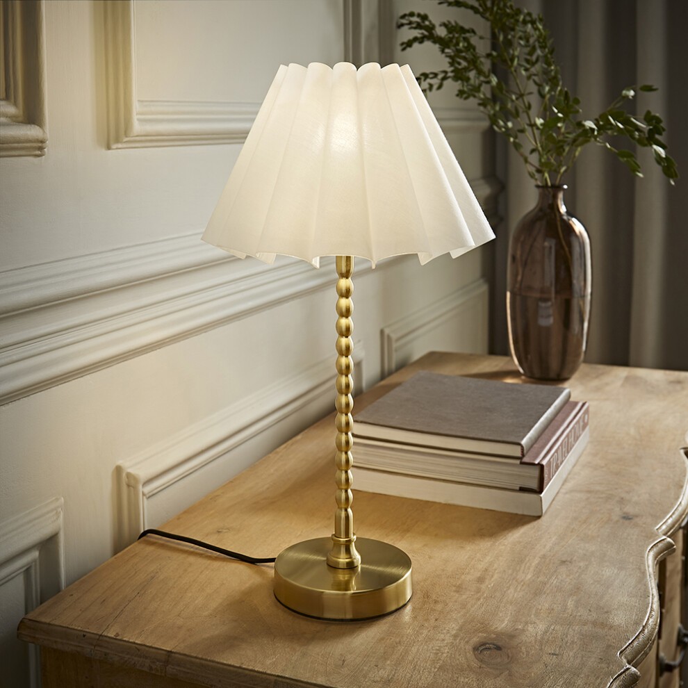Brushed gold bobbin table lamp with a scallop pleated white shade, featuring a sleek design, placed on a wooden surface beside stacked books and a vase with greenery.