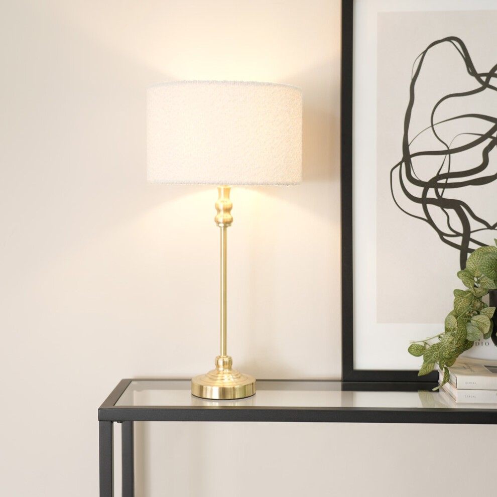 Gold candlestick table lamp with a cream boucle drum shade, featuring a sleek metal base and an LED bulb, placed on a modern black glass table beside a framed abstract artwork.