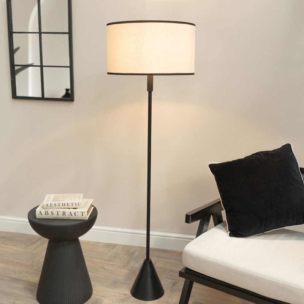 Natural linen trim lamp shade atop a black wood triangle floor lamp, featuring a sleek design, LED bulb, and a cozy atmosphere, complemented by a minimalist side table and cushion.