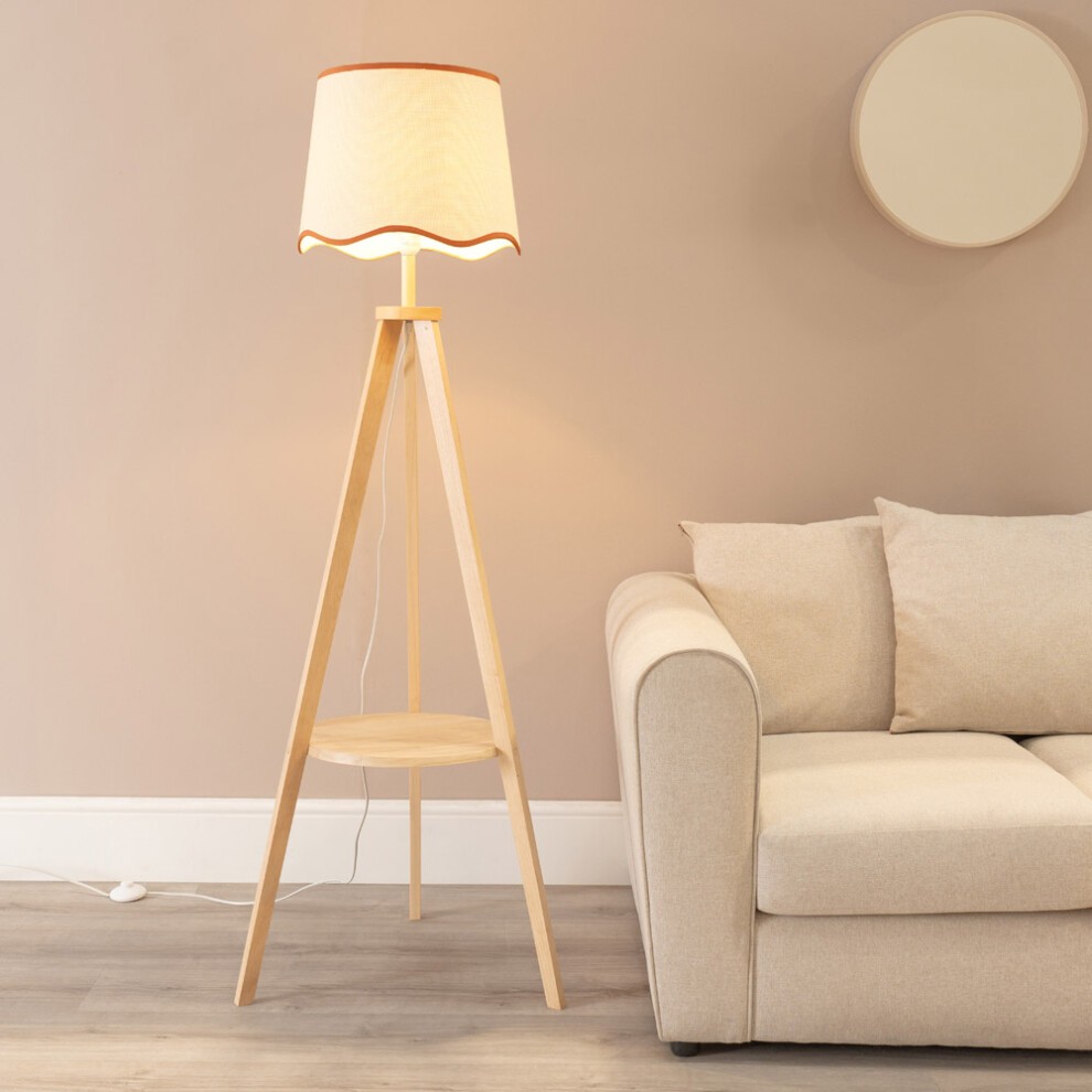 Natural wood tripod floor lamp with a scalloped tapered shade, featuring a warm light bulb, set beside a light beige sofa against a soft, neutral wall.
